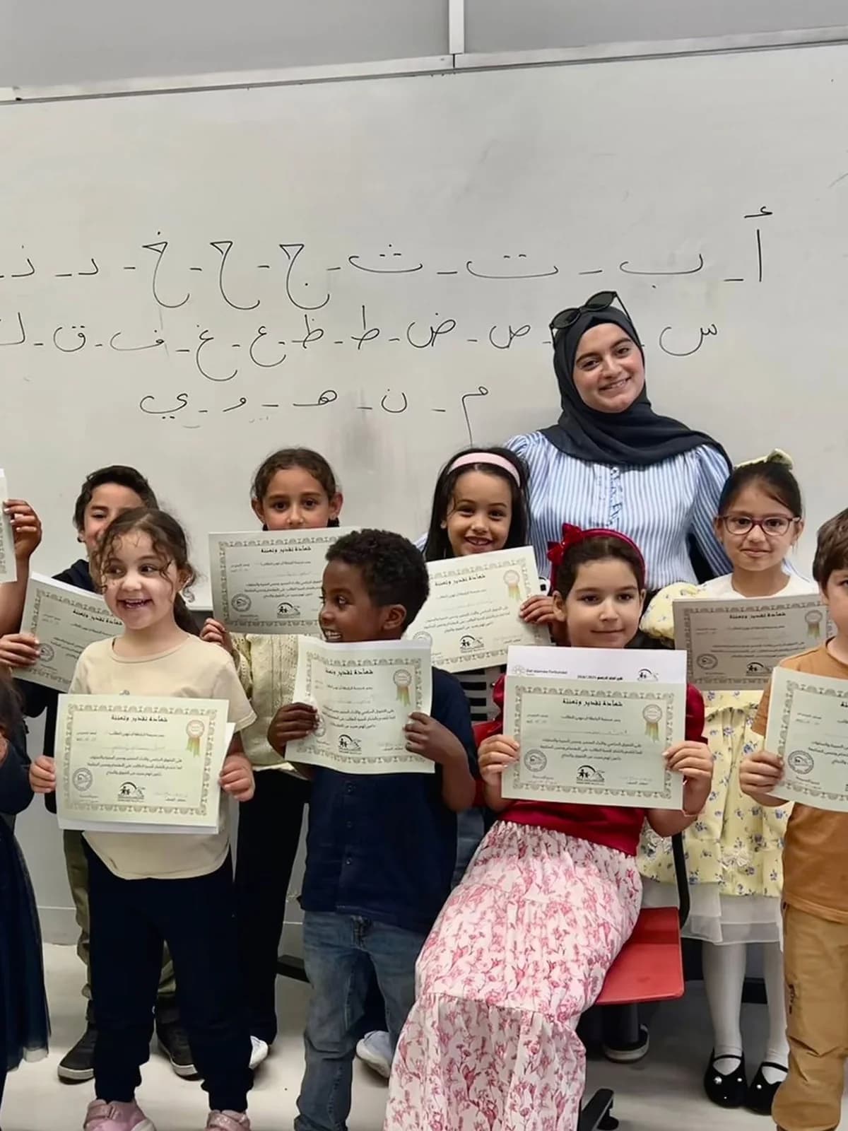 Weekend school at Rabita: pupils holding their certificates with their teacher, the Arabic alphabet on the board behind them