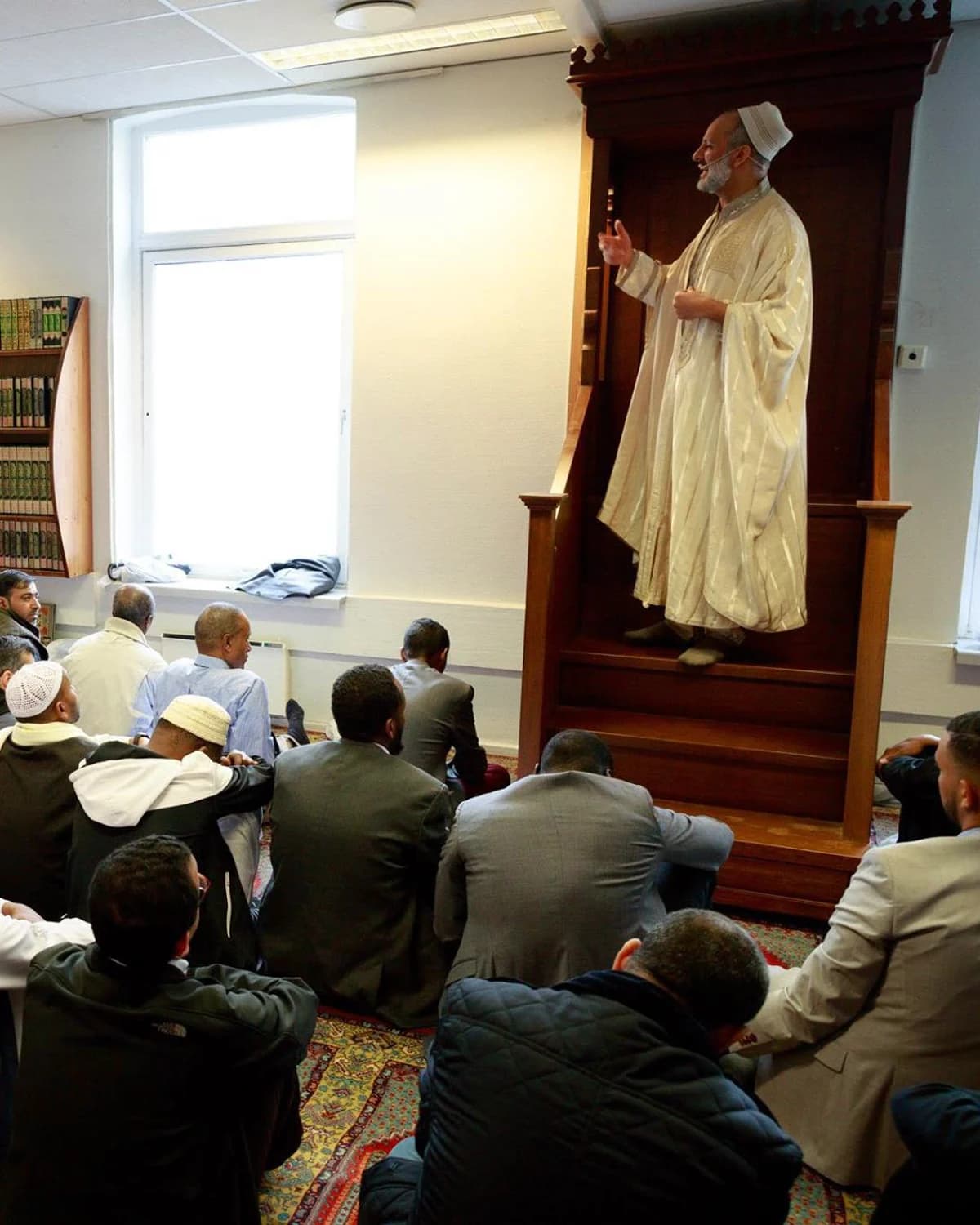 The Friday sermon in the old prayer hall: an imam on the minbar, the congregation seated on the carpet below